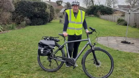 James is wearing a his vis yellow waistcoat and w white bike helmet. He is holding a dark grey e-bike and in standing on the lawn in a garden.
A mixed hedge is on the left and a fence is on he right of the picture.