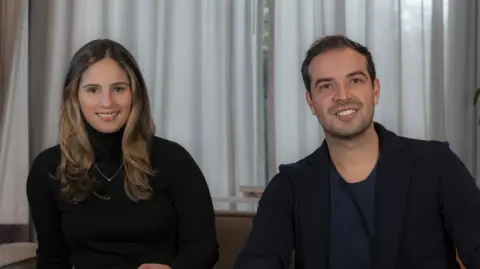 Foodology Smiling, the Foodology founders sit next to each other. Daniela Izquierdo wears a black turtle-neck jumper with a fine chain and Juan Guillermo Azuero wears a navy blue crew-neck shirt and black gilet.