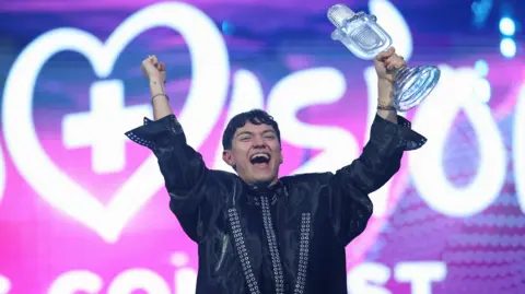 Reuters JJ holding a glass microphone trophy aloft in celebration with his other hand in the air against a purple and pink background 