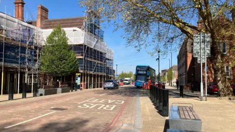 A stretch of pink coloured paved road, with the words bus gate on it, and a bus parked up in the distance. There are buildings to the left and right of the road