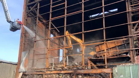 Firefighter uses water hose at the damaged site. The building is almost reduced to rubble with only a metal frame holding one side of it up.