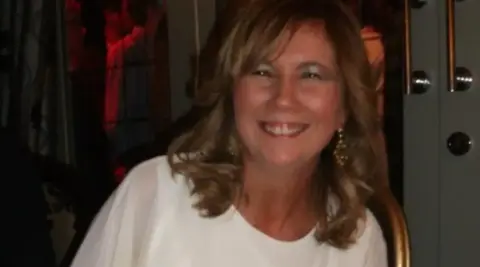 A woman with long hair who is wearing a white top is smiling at the camera