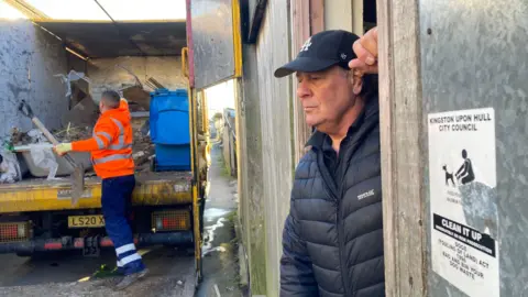 Jo Makel / BBC A casual side profile of Darren Speck, wearing a black cap and a black padded Regatta coat. He is leaning against a door frame with his left hand. In the background is a man in blue trousers, black boots and an orange hi-vis jacket putting the rubbish into a yellow van. 