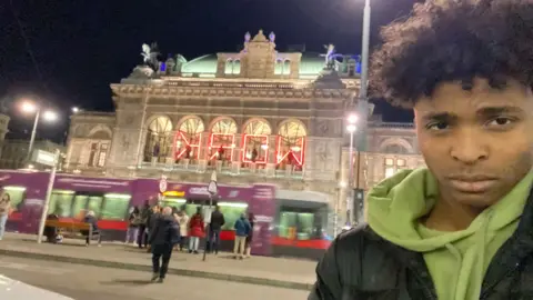 Luka Chijutomi-Ghosh Luka fica ao lado de um prédio em Viena, com um bonde passando atrás dele. Ele está olhando para a câmera. 