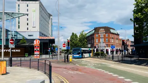 A blue and white bus navigating a road junction. There is a red-coloured bus lane and a green-coloured cycle lane, with no entry signs and black railings to one side. 
