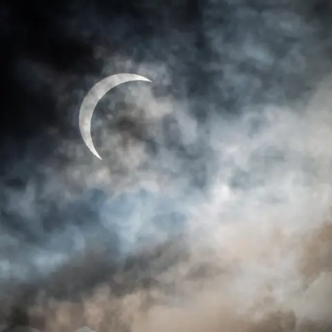 Anastasiia Petruk A thin, bright crescent of the Sun shines through layers of dark grey and blue clouds. The scene has a dramatic, moody look, with dense cloud banks along the bottom of the frame.