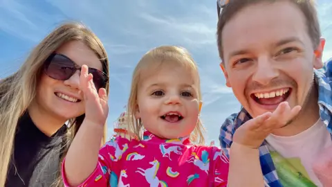 Chris Tuczemskyi A woman and a man posing for a photo with a smiling, female toddler between them. The toddler has her hands raised in the air. The woman is wearing sunglasses. They are set against a bright blue sky.