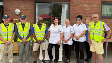 Dennis Briggs Four men wearing yellow hi vis jackets that say "prostate cancer" on them. There are three women, three are in white nurses tops and one in a blue top. they are standing outside of a building and glass double doors