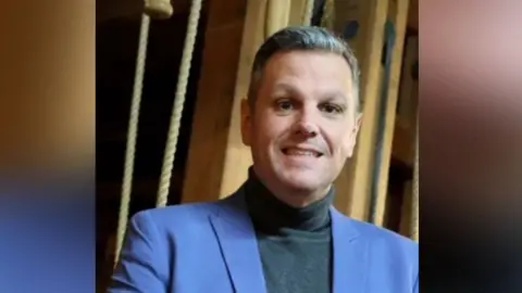 Jonathan Higgins is standing backstage, surrounded by rigging, and smiling at the camera. He has short brown/grey hair and and is wearing a blue suit.