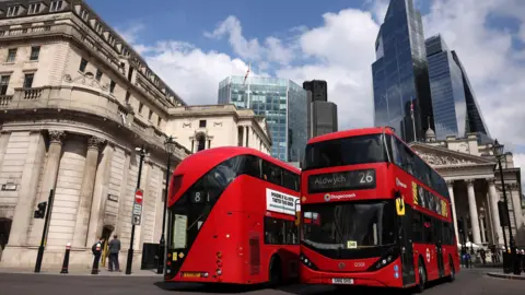 EPA Two red London buses pass on a road in the City of London. Old buildings and skyscrapers are visible against a clear blue sky.