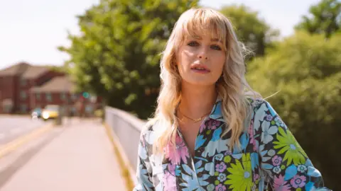 Jess Davies wears a colourful floral boiler suit. She has wavy blonde hair with a fringe. She leans against a railing, with the road in the background blurred. 