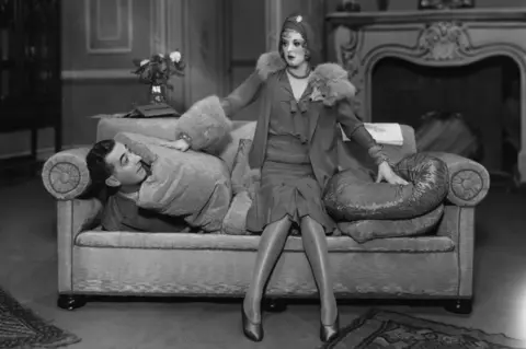 Getty Images Black and white photo of a man beneath cushions on a sofa and a woman sitting on top of the sofa on the stage of Streatham Hill Theatre
