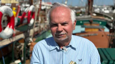 Jodie Halford/BBC A man with white hair and a short, white goatee beard looks at the camera. He is wearing a blue pinstriped shirt with a small black logo on one side. He is standing on a barge.