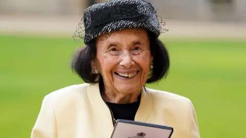 PA Media Lily after being made a MBE at Windsor Castle in January 2023. She has short, black hair and is smiling. She is wearing a cream jacket and black and silver hat.