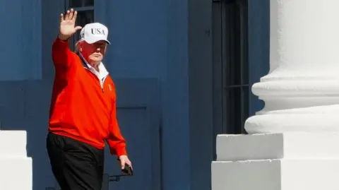 President Donald Trump, wearing a white USA cap and a red golfing sweater, waves as he passes by white pillars outside the White House