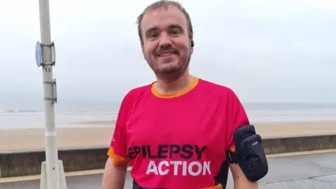 A man wearing a pink sports top smiles for the camera next to the seaside.