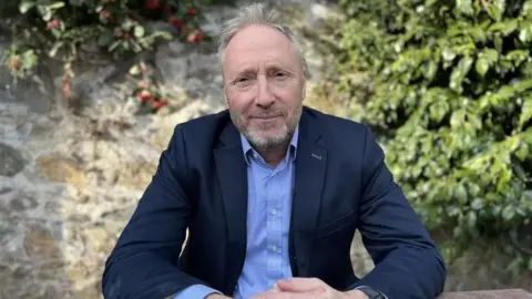 BBC Deputy Paul Montague pictured wearing a navy blazer, and blue shirt. He has short brown and grey hair and beard.