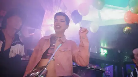 Haus of Fun A female-presenting person dances and sings along to music. They are wearing pink denim jacket, pink metallic crop top and matching cross-body bag.