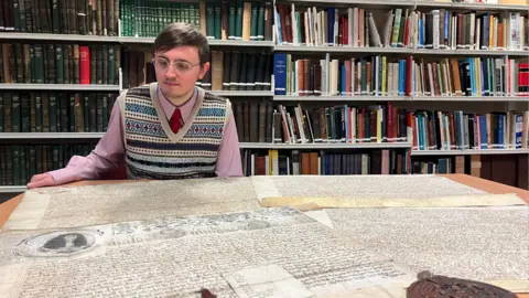 West Yorkshire Archive Service Alex Pearson from West Yorkshire Archive Service is sitting at a table on which are two large vellum Royal Charters.