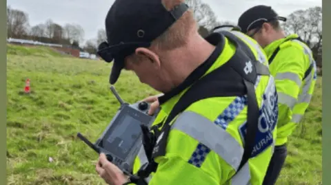 A police officer wearing a yellow hi-vis jacket operating a drone in a field. He is looking at the control screen display.