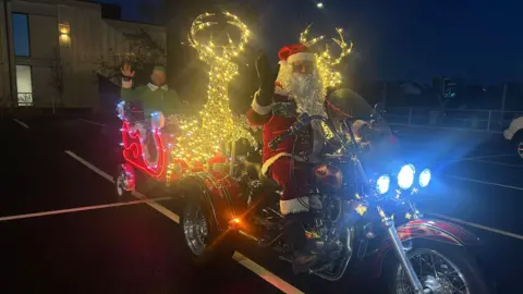 A man dressed in a Santa suit is sat on a red glittery motorcycle with three wheels. Behind him is a sleigh that has been illuminated in red lights with a man in a green elf costume sat inside it. Between the two men are two illuminated golden reindeer. 