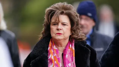 PA Media Elaine C Smith who has shoulder, length brown hair is wearning a pink, orange and white silk scarf and a black fluffy coat. She has a solemn look on her face