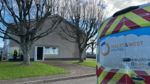 A Wales and West Utilities van parked on a road outside a village hall building. Two trees are planted on the pavement next to the hall. Two people wearing hi-vis jackets are standing outside the building.