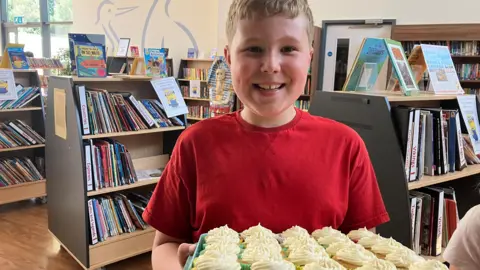 Reuben is standing in a library holding a tray of cupcakes decorated with swirls of white icing. He is wearing a red T-shirt and is smiling at the camera.