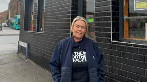 Jonny Humphries/BBC Marie Williams, who has blonde hair and is wearing a blue fleece over a black North Face branded t-shirt, stands in front of Stanleys Cafe which has black tiles installed on the brick-work