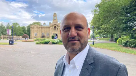 A balding man wearing a grey suit jacket and white, open-necked shirt standing in the park with a Yorkshire stone building in the background.