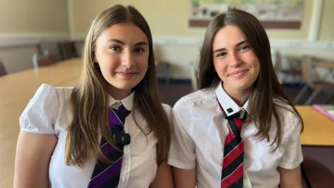 Two teenage girls, both with brown hair, look into the camera. They're in school uniform - white shirts with ties and are sat in a room with school desks in the background.