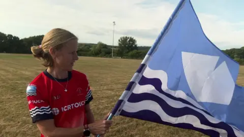 Sammie Mumford standing on a playing field, wearing a red Team Isle of Wight top and holding a blue Isle of Wight flag