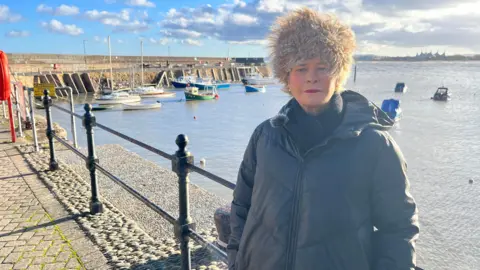 Rachel Gilmour MP is standing by Minehead harbour wearing a coat and fluffy hat. There are small boats in the harbour behind her. She is standing in front of a railing. 