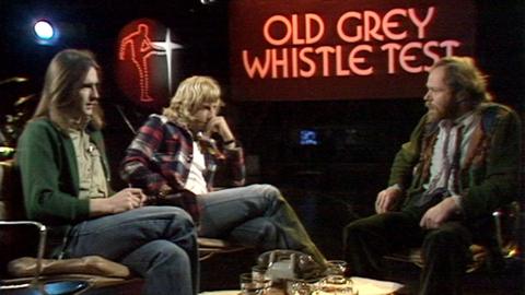Status quo band members sit for interview with presenter in studio. Old Grey Whistle Test signage features on the wall behind them. 
