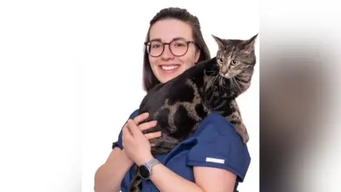 Lime Trees Vets A woman with long brown hair, glasses and a blue vet top smiles as she holds a cat over her left shoulder.