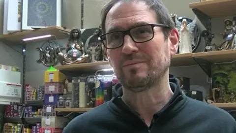 Rocky Salisbury standing in front of a shelf displaying various items, including figurines. He is wearing glasses and a dark top.