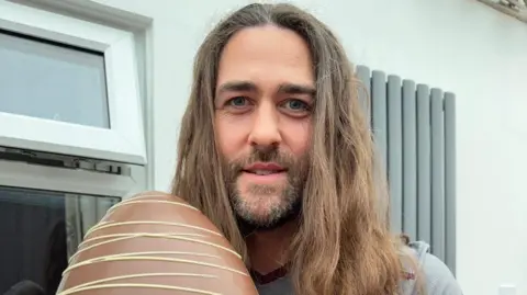 A man with long, shoulder-length brown hair, holding a giant Easter egg