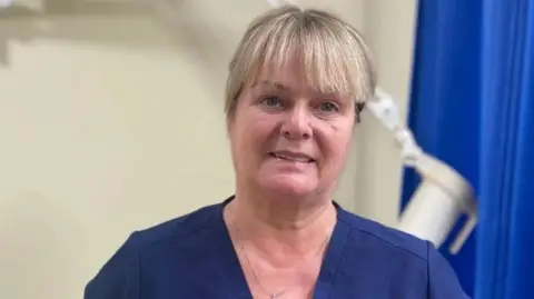 A woman, with blonde hair, wearing dark blue scrubs. 