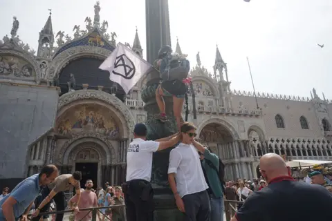 Getty Images Activists protesting in Venice, Italy, against the wedding of Jeff Bezos and Lauren Sanchez. One protester carrying a flag is pictured attempting to climb a column. 