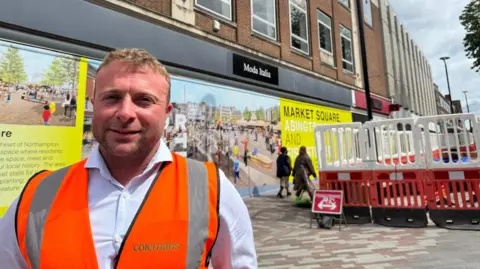 Annabel Amos/BBC Mark Fenning with short blonde hair and a faint beard smiling at the camera wearing an orange hi-vis and white shirt. There is a boarded-up building behind him, showing pictures of the project that is planned for the area. There are barriers separating the public from pavement works behind him.