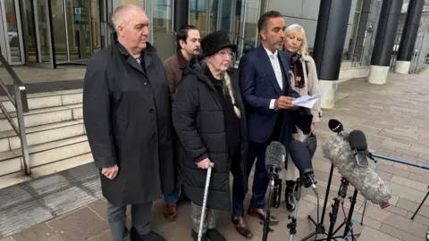 Three men and two women standing in front of microphones, outside a large office building.