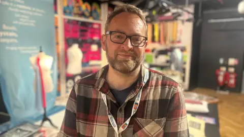 A man wearing a red tartan shirt looks towards the camera. He has black glasses, and a white lanyard around his neck. In the background, a museum is out of focus.