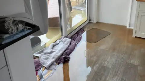 Jan Donald Una casa, con agua dentro, toallas en el suelo, agua en el jardín y un contenedor de basura con ruedas a un lado.