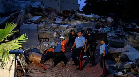 Getty Images An image of six people standing on a pile of rubble, searching for people. They are wearing emergency worker uniforms and four have safety helmets on. The sky is dark.