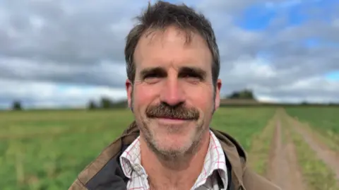 Ben is stood in a field wearing a brown jacket and checked shirt and has a beard and moustache and short brown hair. He is looking directly at the camera and smiling.