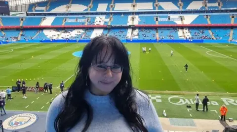 Emily Brobyn Emily Brobyn has long black hair, dark spectacles and is wearing a sky blue sweater. Behind her is the pitch at Manchester City FC with the rest of the stadium in the background.