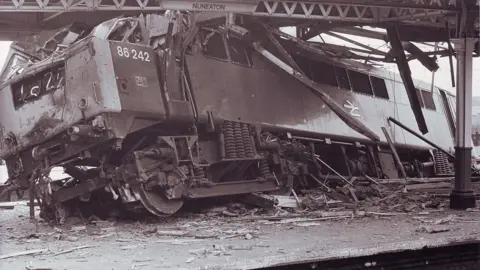 Walter Bloxham/Warwickshire Police/Nuneaton Memories A railway engine at a stop with the front of it on a concrete platform, slightly tilting one side, while the wreckage of the platform roof hangs over the engine which has windows smashed in in this black and white photo.