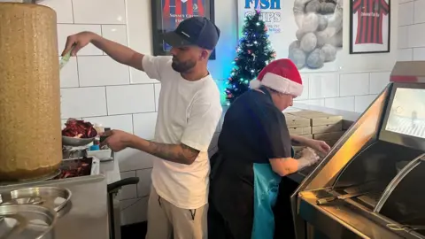 Behind the counter at  Sussex Circus Fish Bar. There are two staff members back to back, a woman looking down at the counter working while they prepare to open and wearing a Christmas hat. A man wearing a cap is working behind her.