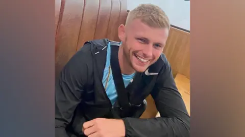 A man with short blonde hair and wearing dark sporting attire with a blue shirt, smiles as he sits at a table and leans on it with his left arm.