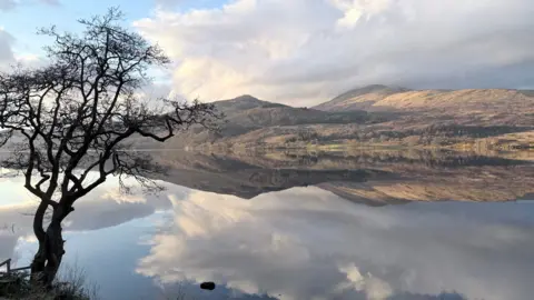 David Strachan A still lake perfectly reflecting the surrounding mountains, clouds, and sky. A leafless tree stands on the left edge of the frame beside the water.